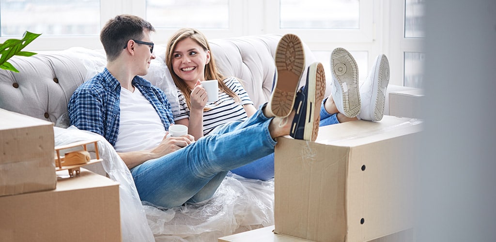 Joyful young couple sitting on sofa covered with plastic wrap, enjoying fragrant cappuccino and chatting animatedly with each other, interior of messy new apartment on background