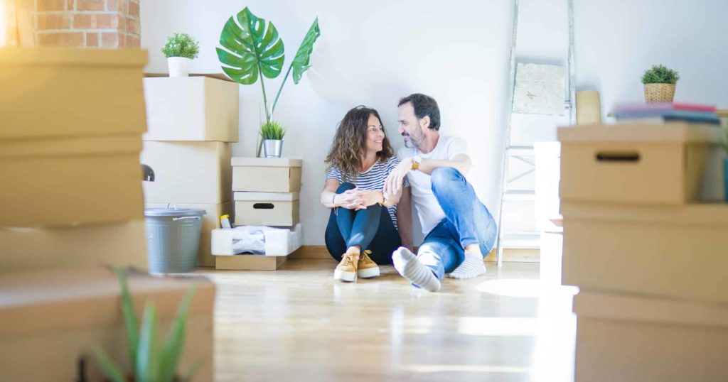 Couple in sitting on the floor with cardboard boxes around and smiling happy for moving to a new home